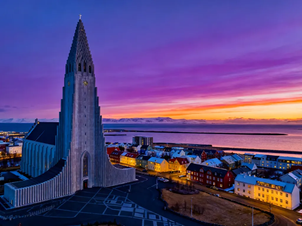 Panorama Reykjavíku o zachodzie słońca z widocznym kościołem Hallgrímskirkja oraz kolorową zabudową miasta nad zatoką.